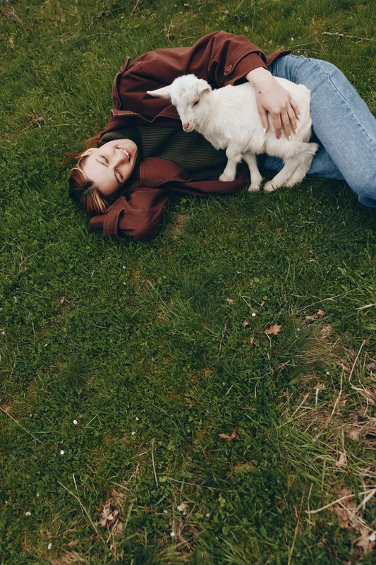 Relaxed lifestyle scene shows a woman lying on grass, cuddling a white kid goat, enjoying a sunny outdoor moment, casual clothing, peaceful rural vibe, warm animal companionship bond