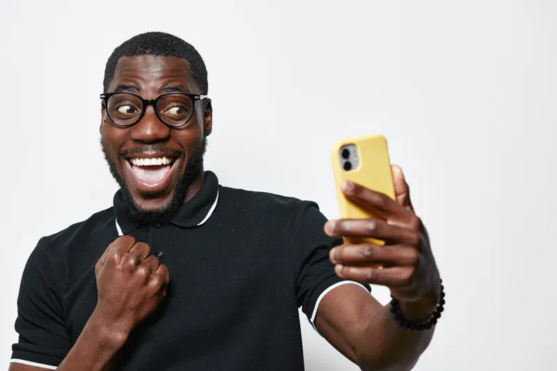 Black man with dark skin and glasses taking selfie with yellow smartphone, smiling and showing excitement in casual black polo shirt on white background.
