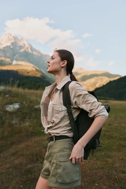 Young woman with brunette hair enjoys a peaceful moment outdoors in casual hiking clothes with a backpack against a mountain landscape at sunset.