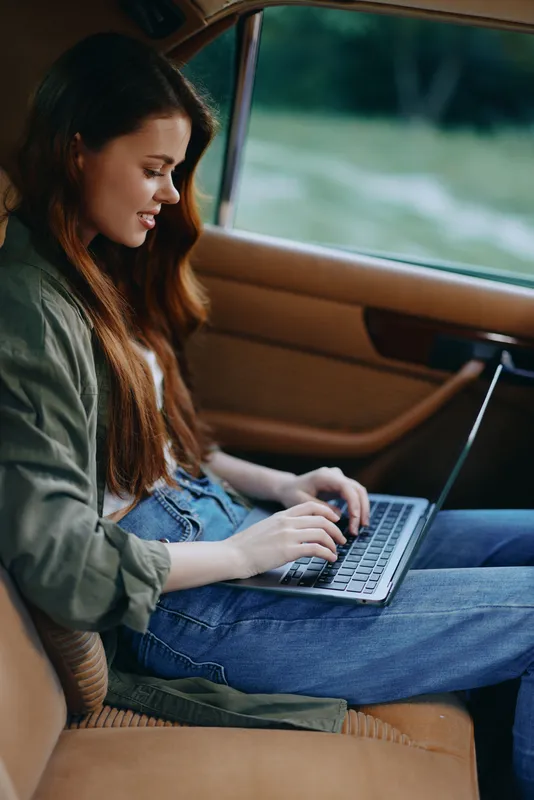 Young woman working on a laptop in a car, wearing casual clothing, with a relaxed expression, surrounded by nature, perfect for remote work concepts.