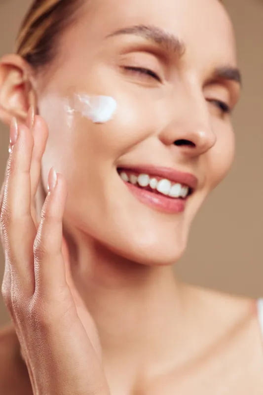 Young woman with clear skin applies moisturizing cream on cheek smiling with closed eyes in soft natural light closeup beauty skincare concept.