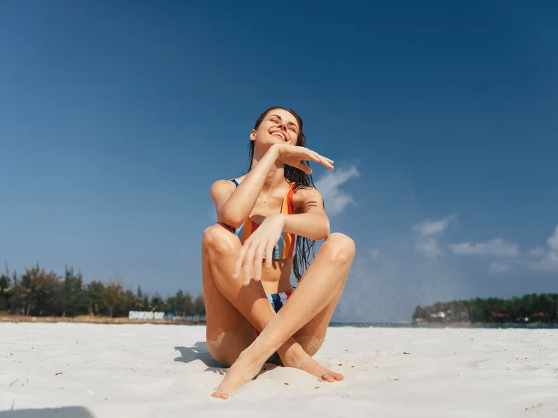 Young woman sits on bright white sand under a clear blue sky, wearing a swimsuit, smiling softly, and enjoying warm sun on relaxed, sunlit skin in a peaceful beach moment.