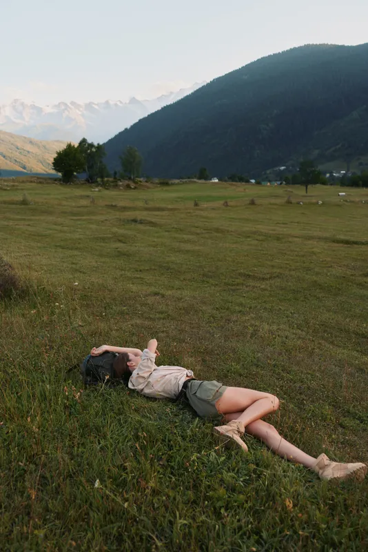 Young woman lying in a grassy field near mountains, enjoying outdoor relaxation, casual clothing, peaceful mood, nature escape, sunlit landscape under a clear blue sky