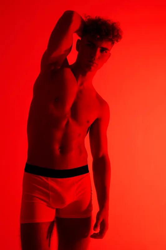 Young man posing topless in red light studio setting with one arm raised behind head wearing boxer briefs for fitness, health, male model fashion, skin and body confident expression.