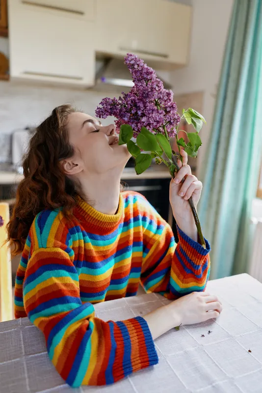 A young woman in a rainbow striped sweater sits at a cozy indoor table, smelling a bouquet of lilac with a calm, joyful expression.