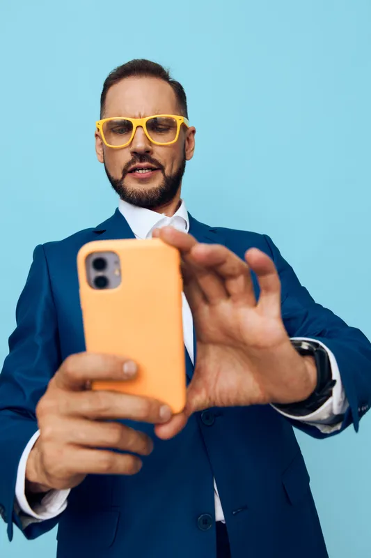 Confident business professional poses for a self portrait against a vivid blue background, wearing a navy suit, yellow glasses, and an orange phone, exuding focus and energetic enthusiasm.