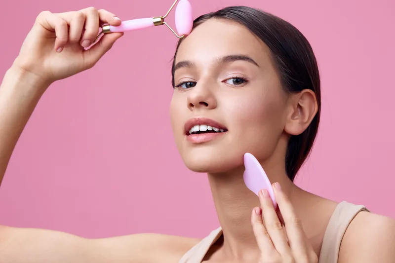 Young woman applying skincare with facial rollers, promoting beauty and wellness, smiling against a pink background showcasing self care routines