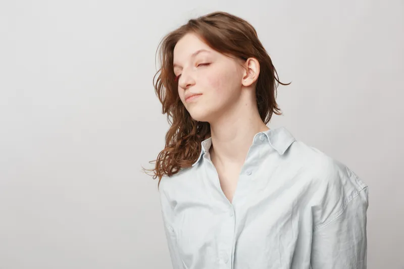 Young woman with closed eyes and curly hair wears light shirt, posing calmly on grey background. Concept of relaxation, peace, and natural beauty in studio portrait.