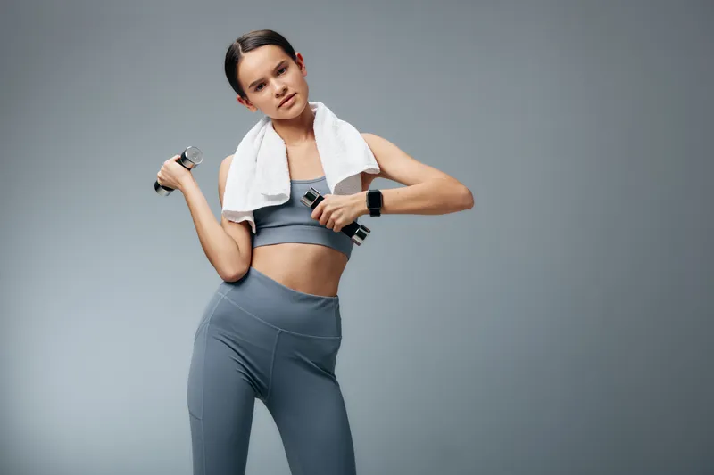 Woman in gray athletic wear lifts dumbbells with a towel over her shoulders, showcasing fitness, sport and workout training in a studio. Focused, strong, healthy energy.