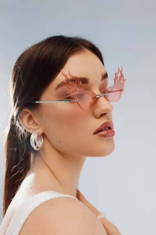 Young woman in unique flame-shaped pink sunglasses poses with a confident expression. Studio portrait on a light background. Concept of beauty, fashion, and style.