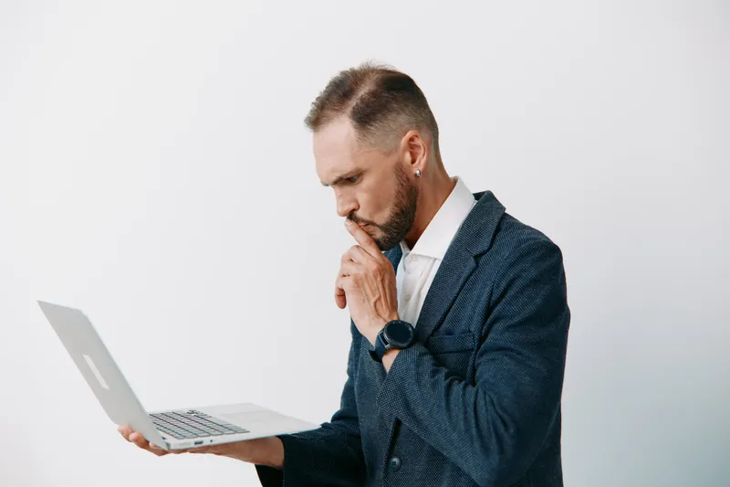 A focused business man studies a laptop against an isolated light backdrop. His professional blazer and calm expression convey concentration, ambition, and careful decision making.