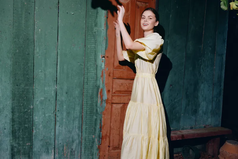 A woman in a yellow dress stands by a wooden doorway, posing with raised arms against a weathered green wall, creating a vintage rustic fashion portrait with soft evening light