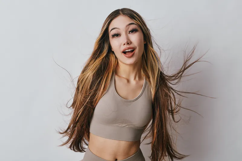 Young woman with long hair smiling and wearing sportswear posing on light background. Attractive female model showing natural expression with casual style and fitness outfit.