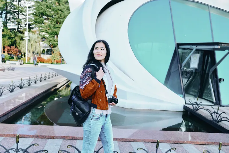 Woman with backpack and camera strolling past futuristic architecture in an outdoor urban setting, fashionably dressed in denim jeans, jacket and scarf, capturing a moment of discovery near glass