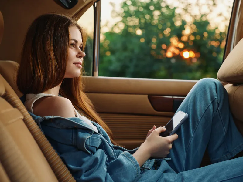 Young woman in denim outfit sitting in a car, holding a phone, enjoying a sunset view with a calm expression, capturing moments of tranquility on a journey.