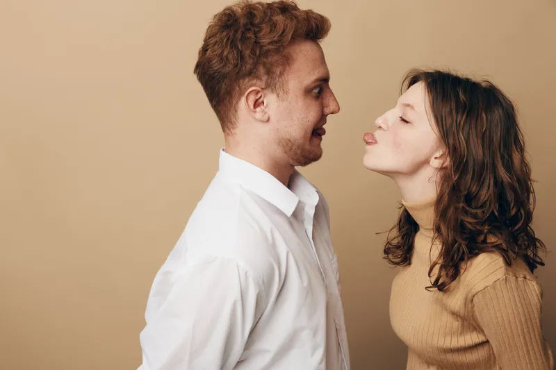 Young woman playfully sticks out tongue at smiling man in white shirt. Casual interaction between couple on beige background. Concept of fun, relationship, and lighthearted moment.