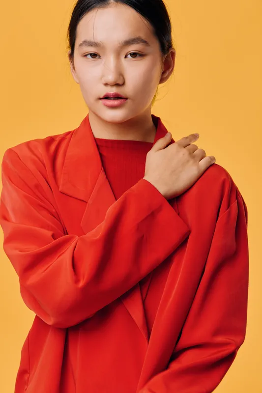 Young woman posing in bright orange oversized jacket against yellow background with calm expression and natural makeup, fashion portrait with styled hair and casual look.