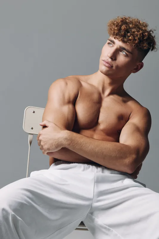 Young man with curly hair shirtless sitting on chair with crossed arms, posing and looking side with serious expression against gray background. Fit model with strong muscles and white pants.