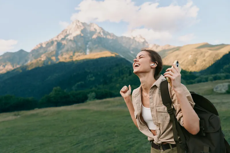 A joyful woman hiking outdoors with a backpack smiles on a grassy hillside, with towering mountains in the background, capturing a moment of wanderlust.