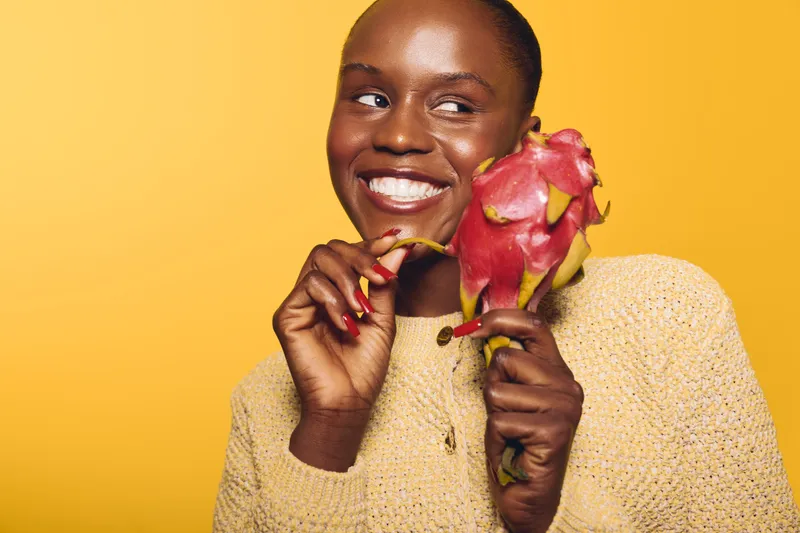 Smiling black woman with dark skin holds a dragon fruit near her face, wearing a beige sweater against yellow background. Concept of healthy lifestyle, natural food, and positive mood.