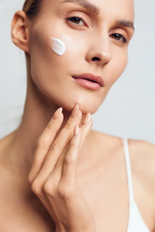Close-up of a young woman applying white face cream on her cheek. Smooth skin and natural makeup emphasize skincare and beauty routine with soft lighting on a plain background.