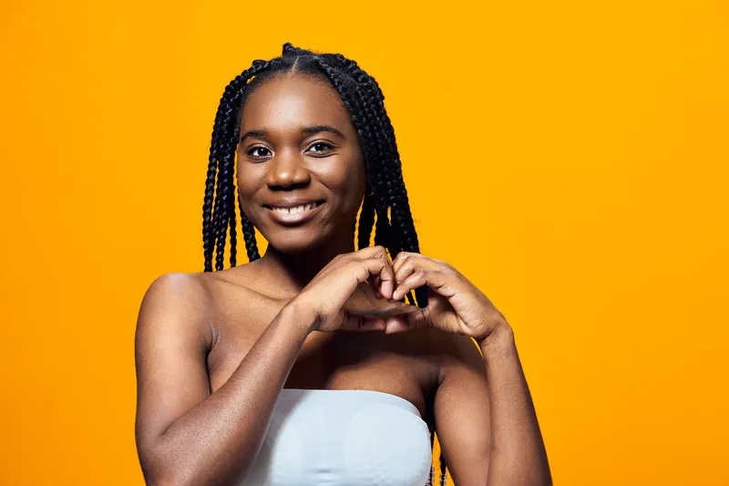 Smiling young Black woman with braided hair showing heart gesture against a bright yellow background, promoting love and positivity in a vibrant seasonal concept