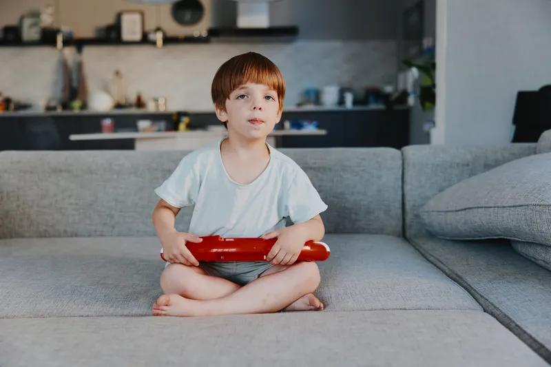 Happy boy sitting on a couch, playing with a red toy, enjoying indoor time in a cozy home setting, wearing casual light clothing, relaxed atmosphere.