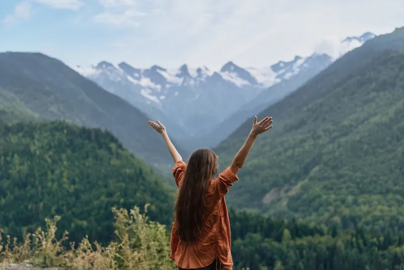 Woman standing with arms raised outdoors amid a vast nature landscape, gazing toward snowy mountains and lush valley, expressing freedom and awe in a scenic travel moment.