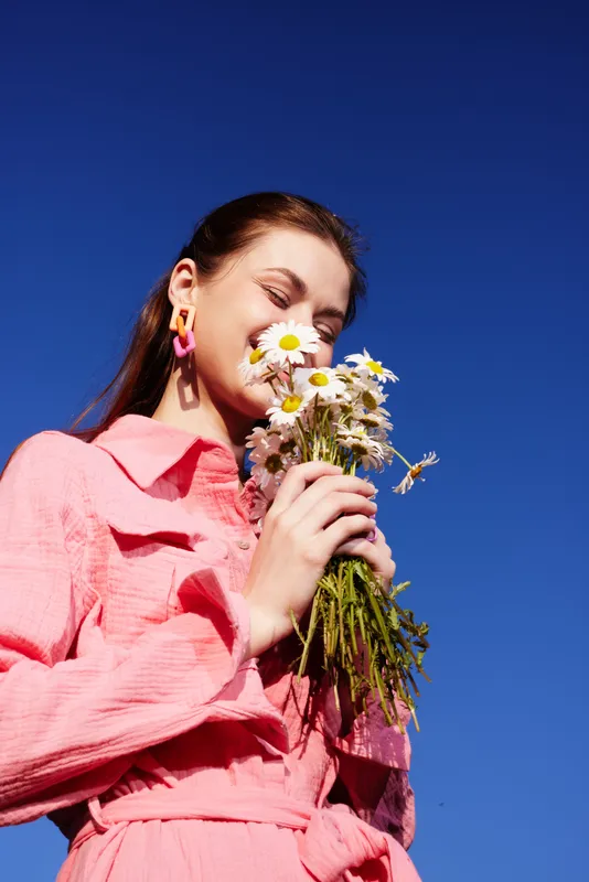 Young woman in pink dress joyfully holding flowers against a bright blue sky, embodying happiness and spring vibes with a cheerful demeanor