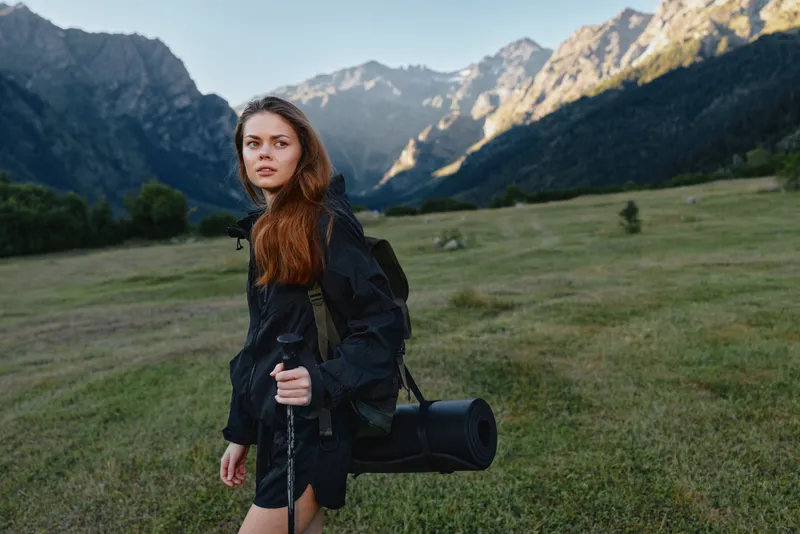 A lone female traveler stands in a wide open meadow, wearing a dark rain jacket and carrying a rolled mat. She looks ahead with calm confidence, ready for an outdoor journey.