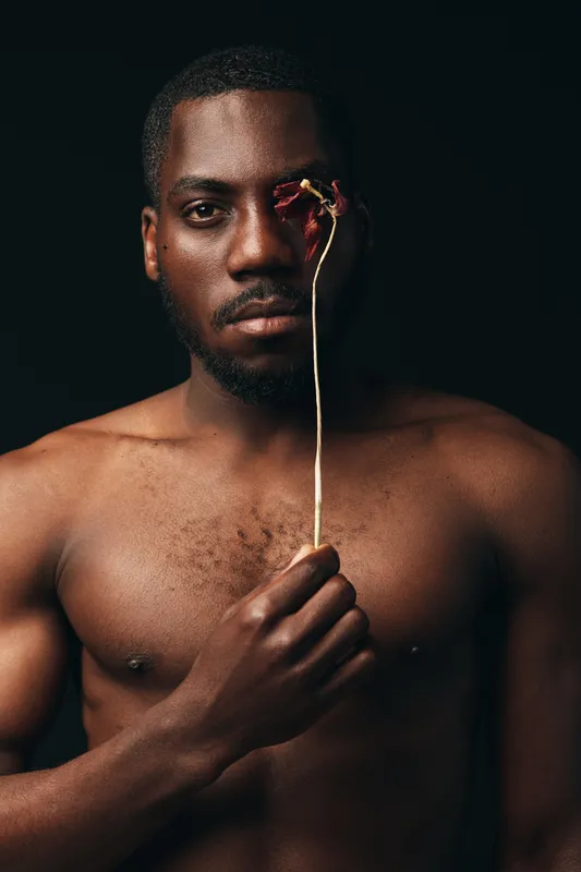 Creative conceptual portrait of African man with dark skin holding dried flower covering one eye, artistic visual style with emotional expression and minimalistic black background.