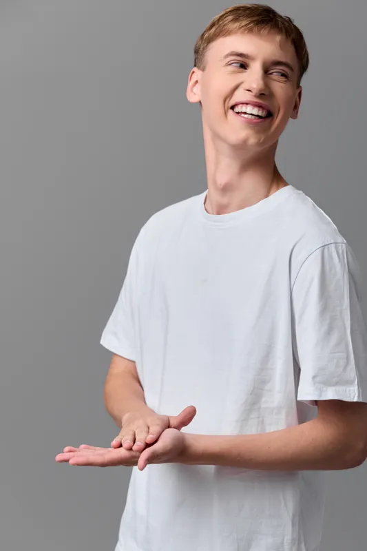 Smiling young man in a white tee stands against a neutral backdrop with open hands, conveying friendliness, approachability, and casual everyday positivity