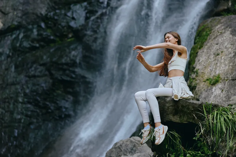 young woman enjoying waterfall outdoors, white activewear, relaxed mood, natural setting, healthy lifestyle, serene atmosphere