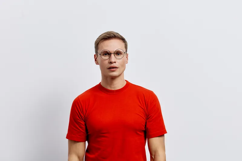 young caucasian man wear red t-shirt and round glasses stand isolated on gray background look camera serious facial expression concept cyber security digital nomad technology