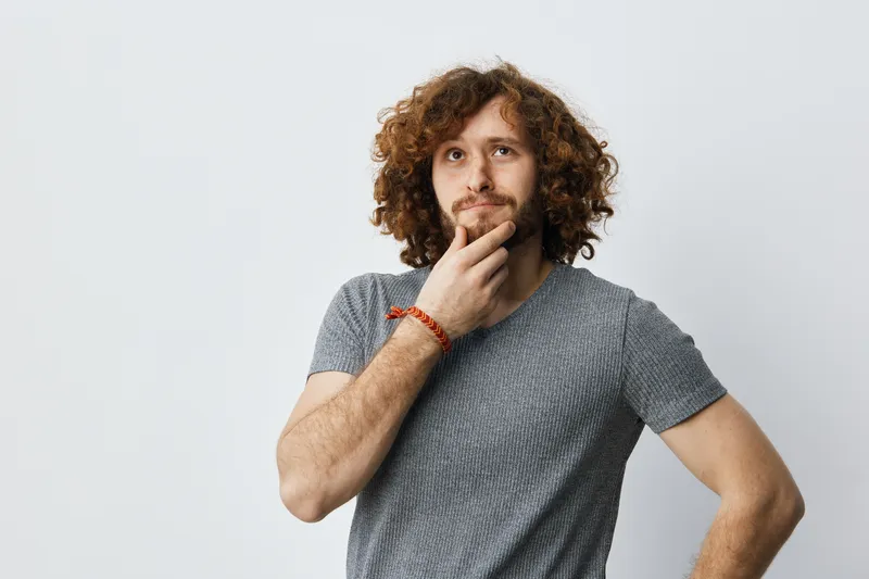 Thoughtful young man with curly hair contemplating ideas against a minimalistic background, showcasing a calm demeanor and casual attire