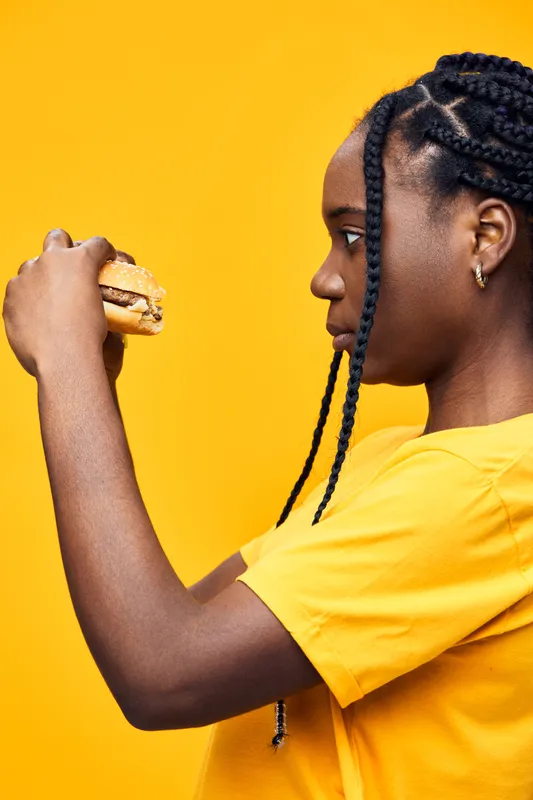 A young African woman in a yellow shirt thoughtfully holds a burger, showcasing healthy eating habits and vibrant emotions against a bright yellow background