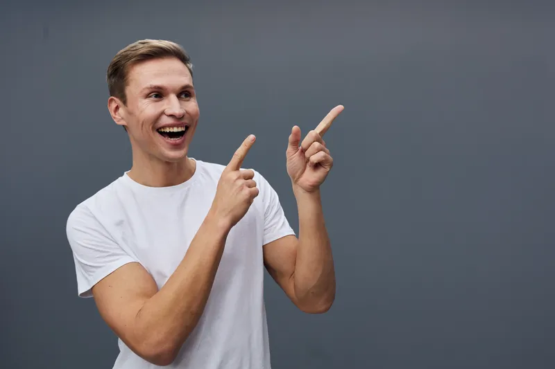 Smiling man with short brown hair wearing a white T-shirt, pointing to his right with both hands and looking cheerful, studio shot on plain gray background, casual lifestyle concept.