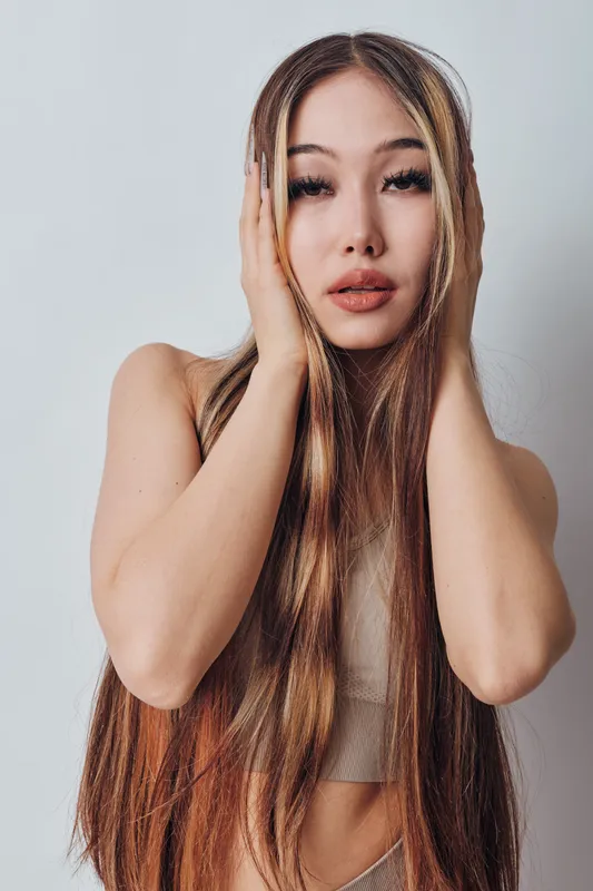 Woman with long brown hair and natural makeup posing with hands on face against plain background. Beauty portrait of young female model with smooth skin and expressive eyes.