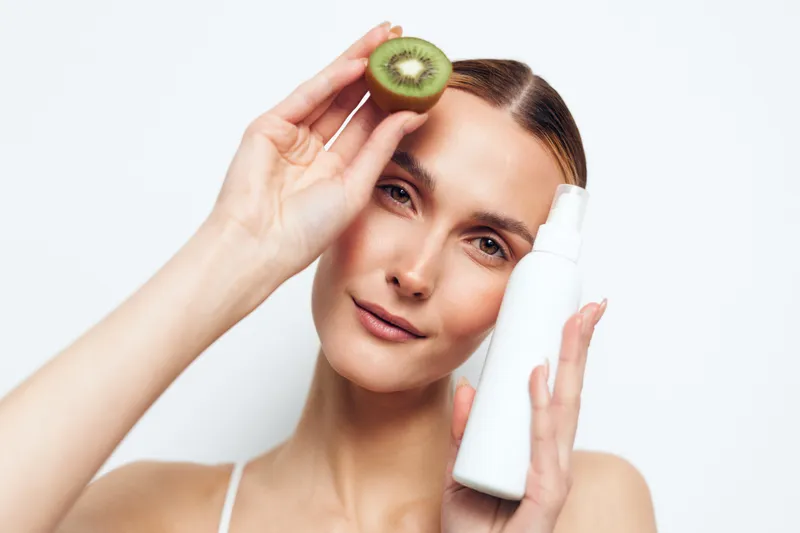 A young woman holds a sliced kiwi and a white cosmetic spray bottle against a white background. The image represents natural skincare and beauty product concepts.