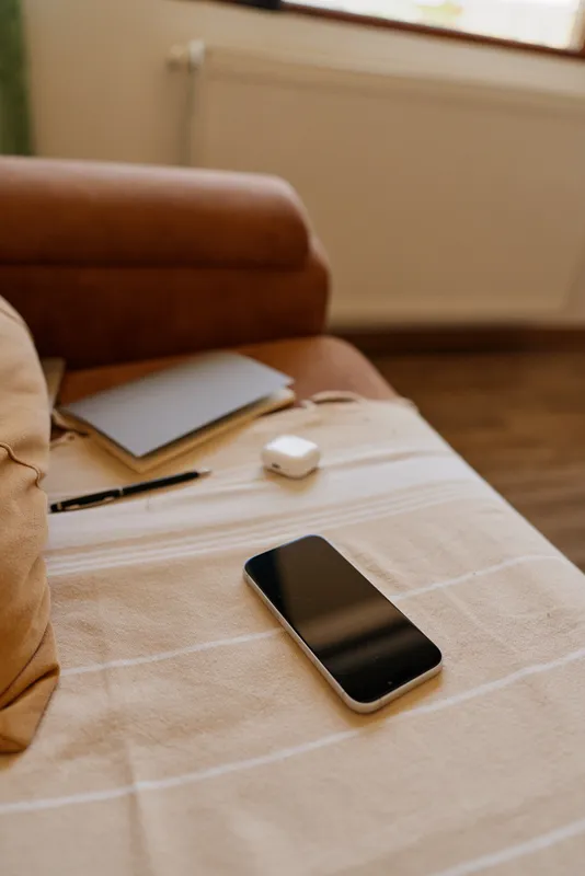 Calm home scene with a smartphone resting on a bed beside a notebook, inviting a relaxed lifestyle moment, soft light, cozy textiles, simple modern interior, quiet morning ambiance