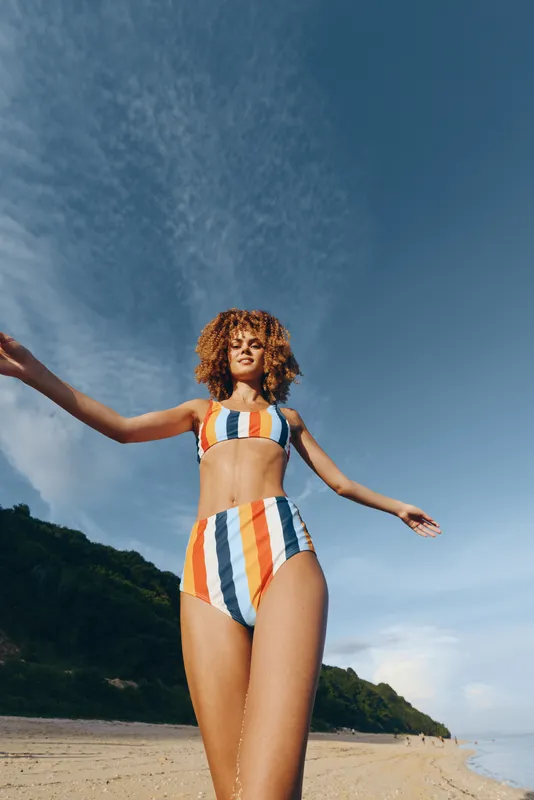 Woman in bikini on beach, summer vacation, curly hair, arms open wide, joyful pose under blue sky, sunny day on sand, outdoor lifestyle, carefree mood, ocean shoreline, peaceful atmosphere