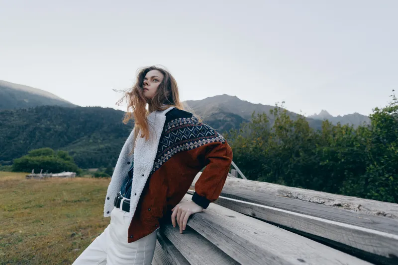 Woman fashion portrait outdoors in mountains wearing patterned sweater and coat, leaning on wooden fence with long hair in wind, moody natural light and casual styling.