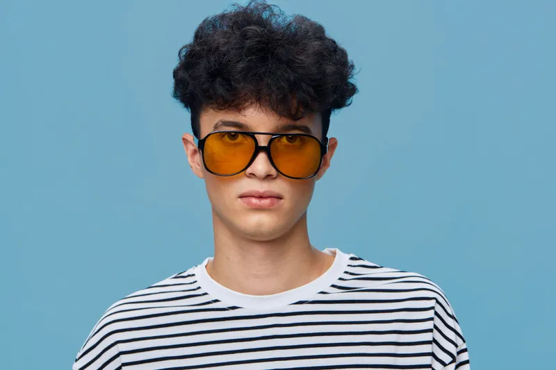 young man wearing orange sunglasses and striped shirt with curly hair standing against a plain blue background in a casual style