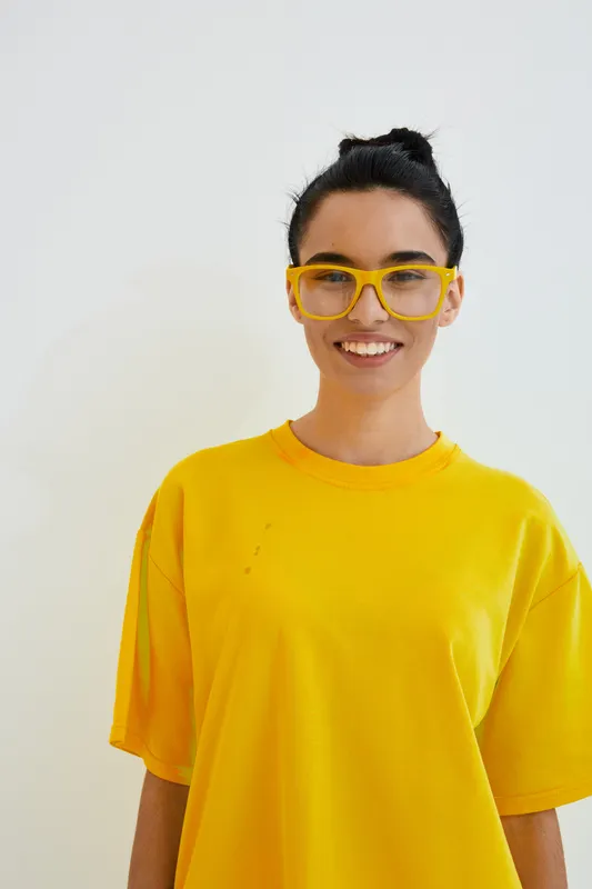 smiling woman wearing oversized yellow glasses and bright yellow shirt stands against plain white background, displaying cheerful expression and casual style.