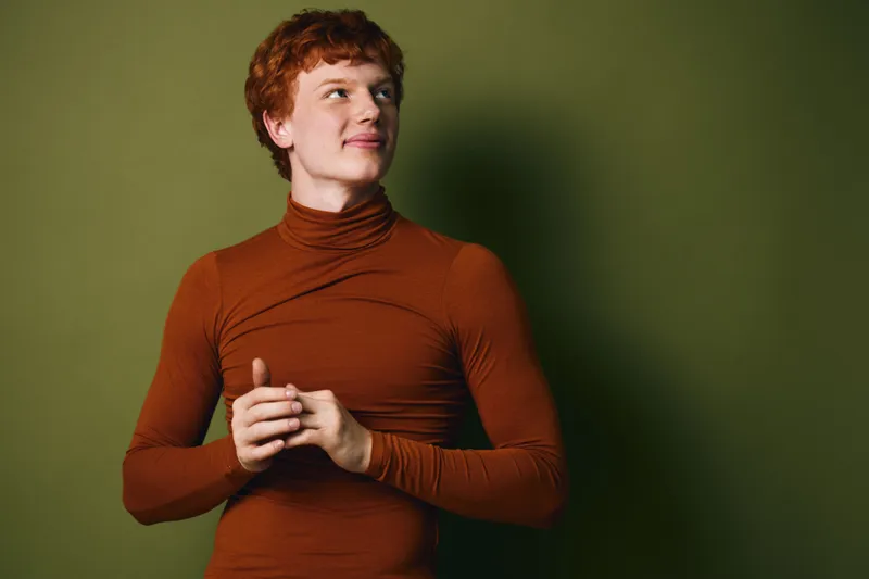Young man with red hair wearing a brown turtleneck sweater standing against green background looking to side with slight smile and relaxed posture in studio portrait.