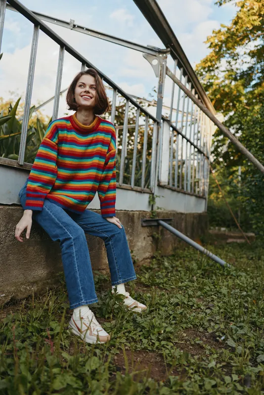Young woman striped sweater greenhouse sitting smiling leaning on concrete ledge in outdoor garden, casual outfit with denim jeans and white sneakers, relaxed lifestyle and nature portrait.