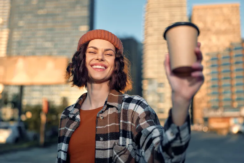 Woman coffee smile city beanie urban portrait: smiling woman holds coffee cup on a city street at golden hour glow, candid lifestyle image highlighting authenticity, mindful living and emotion.