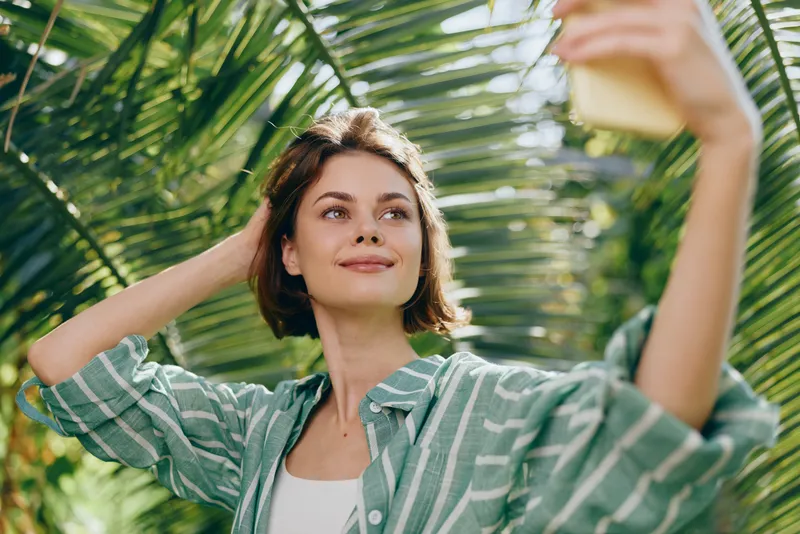 A cheerful woman takes a sunny selfie under lush palm leaves, radiating relaxed confidence and joy. The natural setting showcases brightness, style, and a fresh outdoor mood.