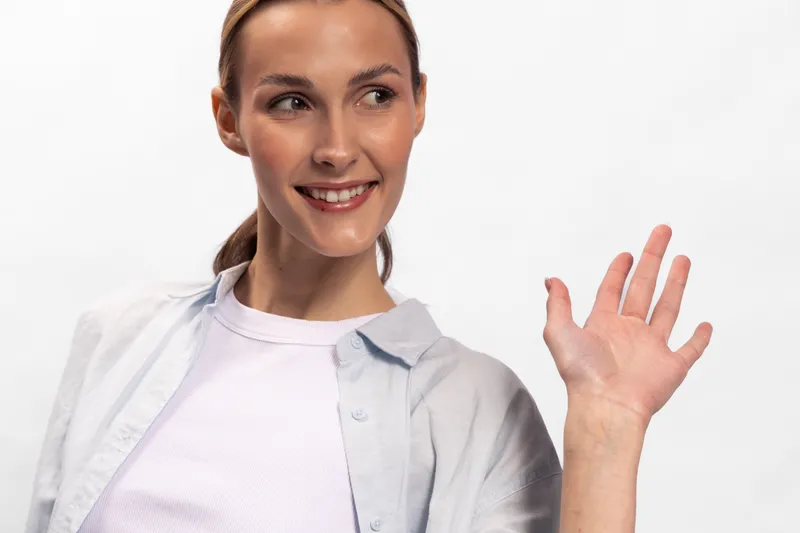 Smiling woman with light skin and blond hair waves her hand in greeting against a white background. Casual clothing and natural makeup create a friendly, approachable mood.