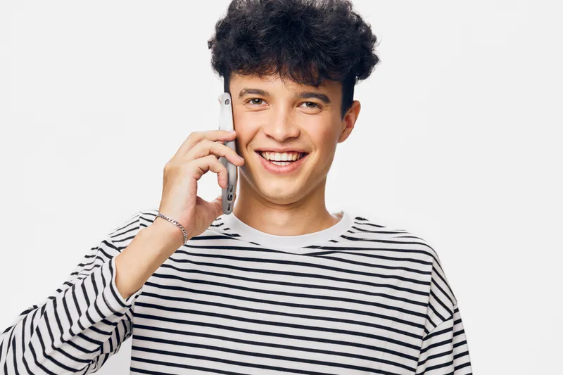smiling man talking on smartphone wearing casual striped shirt with curly hair, white background, happy young adult male communication portrait